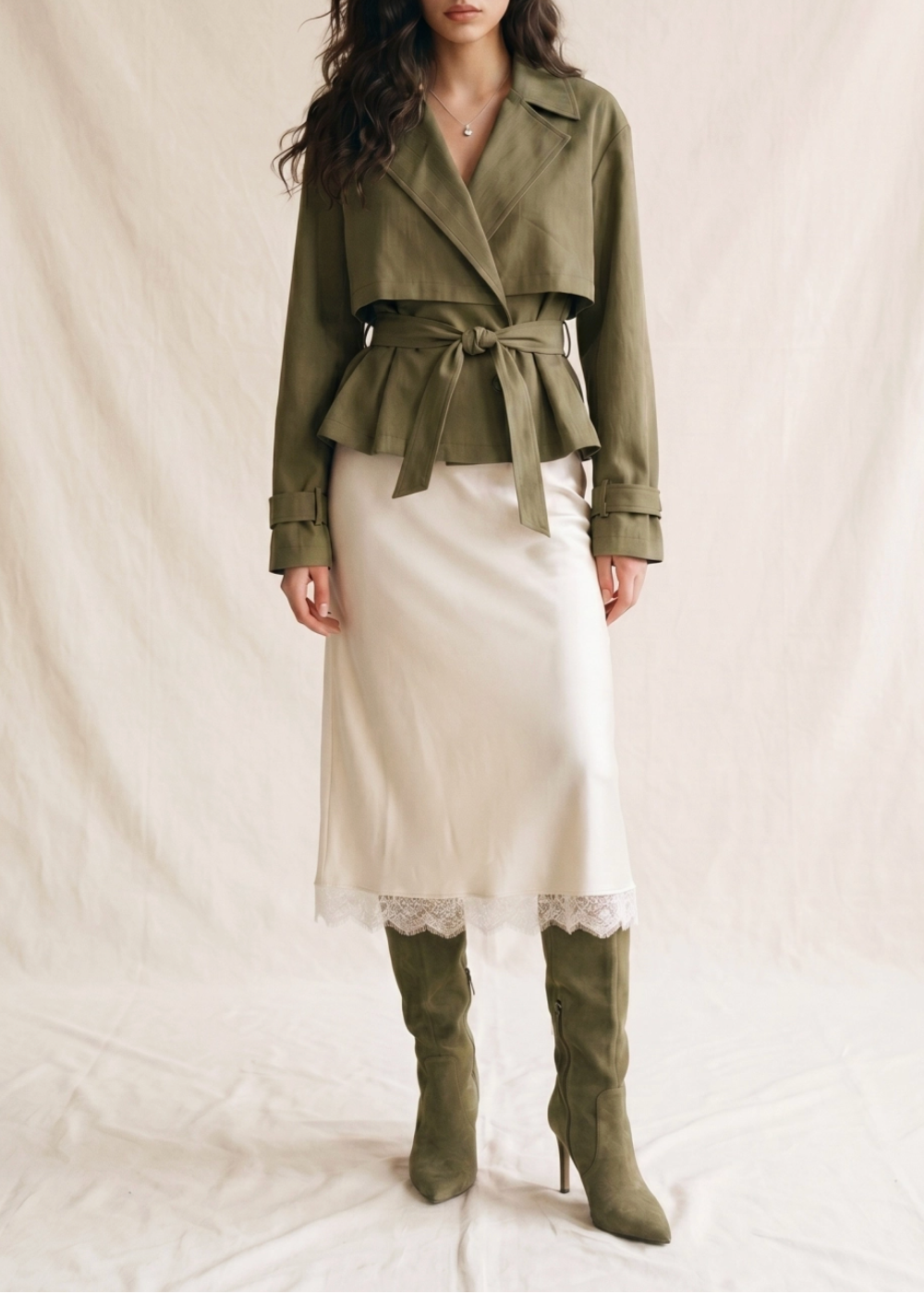 A model wearing an olive green belted wrap jacket, a cream satin midi skirt with lace trim, and matching olive green heeled boots, standing against a draped cream fabric backdrop.
