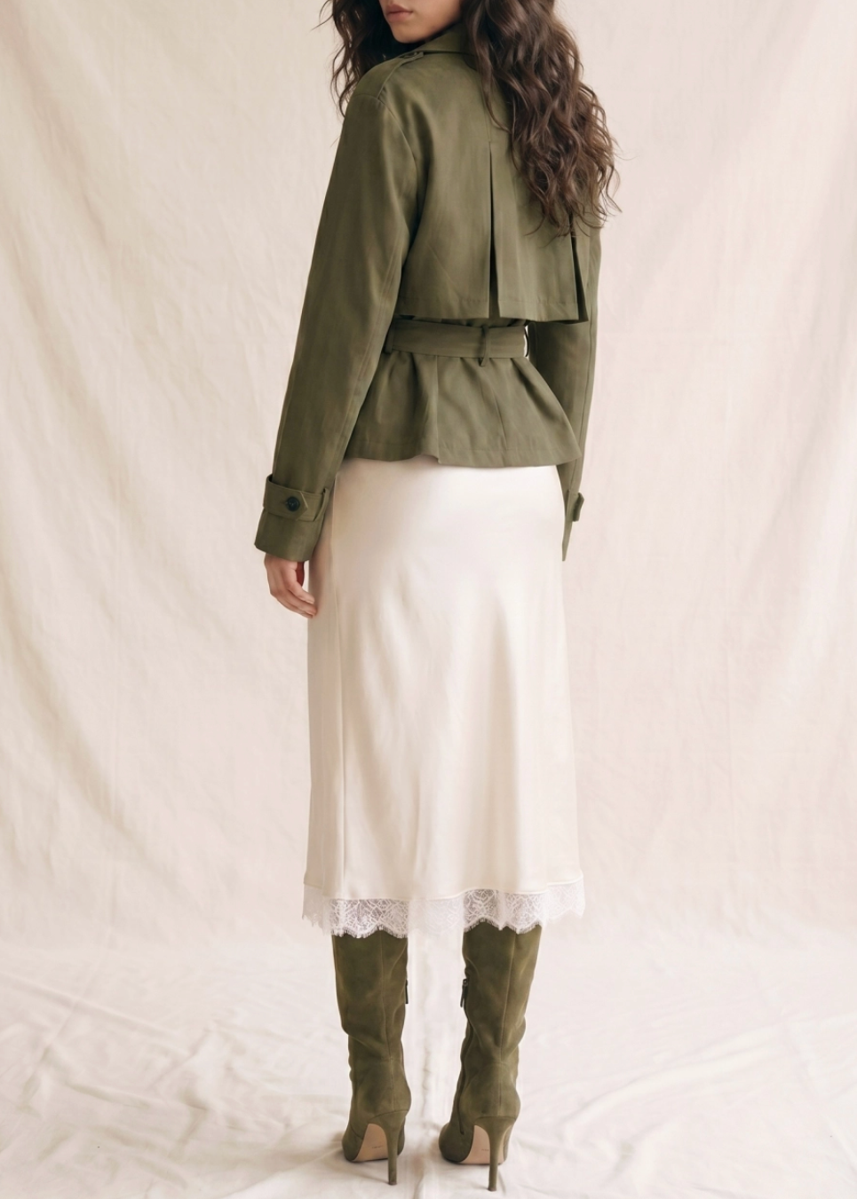 A woman stands with her back to the camera wearing an olive green belted jacket, a white midi skirt with a scalloped lace hem, and olive green knee-high boots, posed against a draped off-white fabric backdrop.
