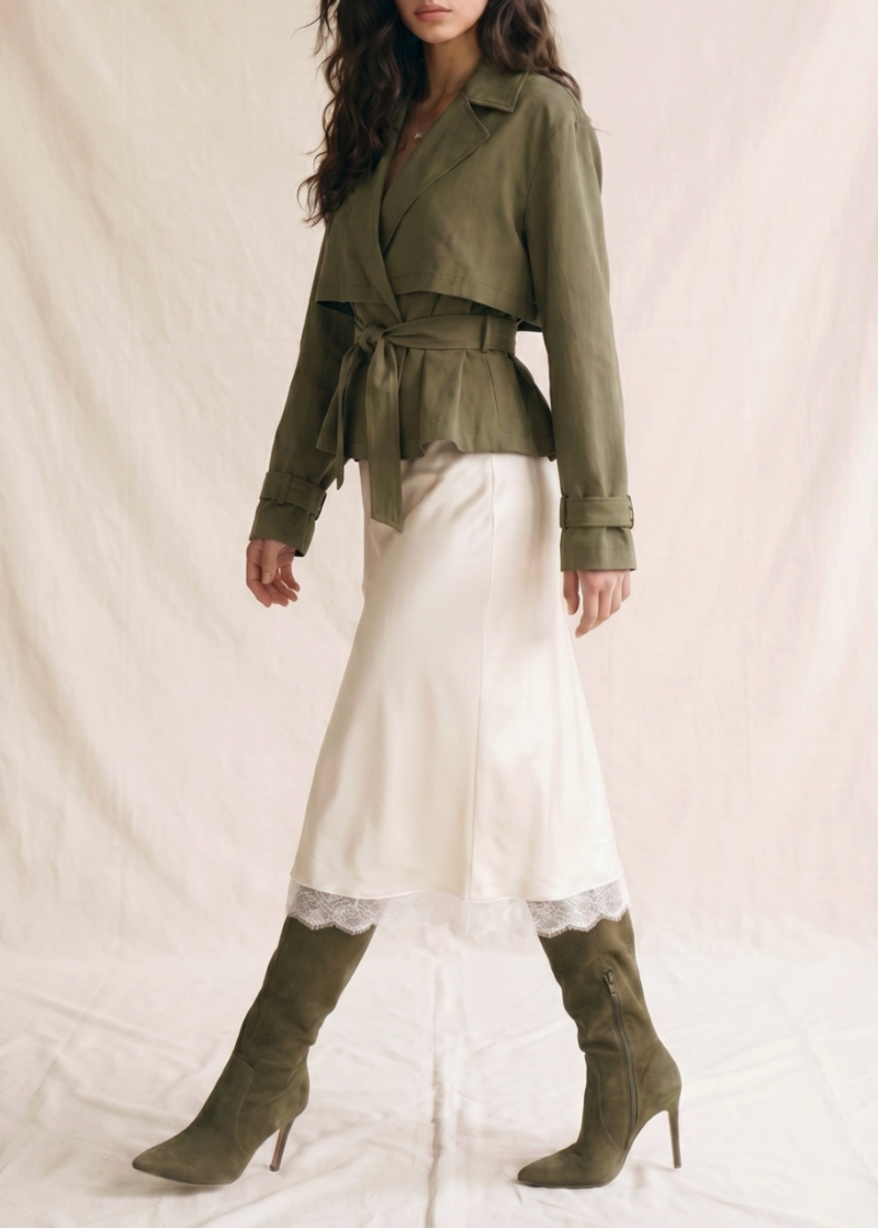 A model wearing an olive green belted wrap jacket, a white satin midi skirt with lace trim, and matching olive green heeled boots, posed against a draped cream fabric backdrop.